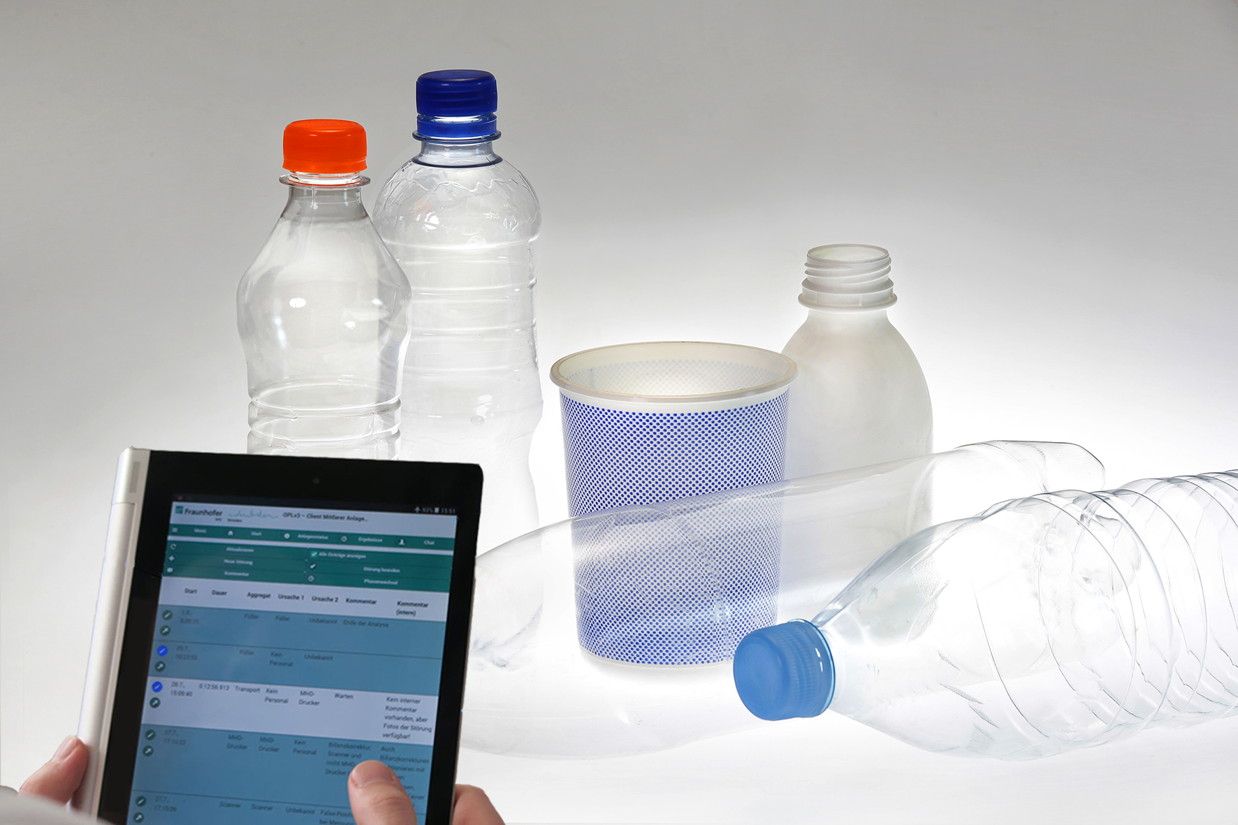 Einige Plastikflaschen stehen in einem weißen Raum, im Vordergrund bedient eine Person ein Tablet, welches Daten zu verarbeiten scheint. (C) Fraunhofer IVV