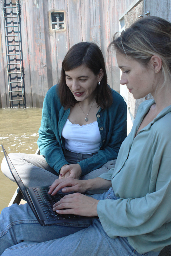 Zwei Personen blicken gemeinsam konzentriert auf einen Rechner. Sie sitzen an der Brüstung des Hamburger Hafens, im Hinztergrund ist grünliches Wasser und eine Leiter zu sehen.